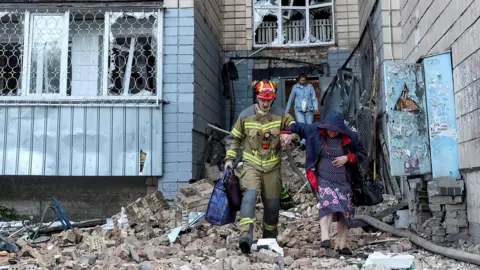 A firefighter helps an old lady out a building holding her arm as they step over piles of bricks and rubble