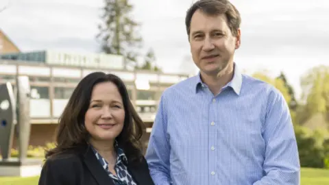 The Natasha Allergy Research Foundation A photo of Tanya Ednan-Laperouse, a woman with brown hair wearing a black jacket and blue and white shirt, stood next to her husband Nadim Ednan-Laperouse, who is wearing a blue shirt. There is a building and greenery behind them.