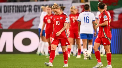 Jess Fishlock walks forward with her head bowed after an England goal. She is in a red Wales kit. Ffion Morgan is to her left, with head turned behind to the England and Wales players out of focus in the background. Wales flags are draped over sponsorship boards.