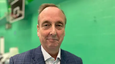 BBC Ian Thorn smiles at the camera. He is wearing a blue jacket and check shirt. Behind him is a green wall