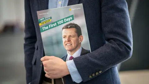 Rob Norman/Plaid Cymru A man holding a report that says "the first 100 days" with a picture of Rhun ap Iorwerth's face.