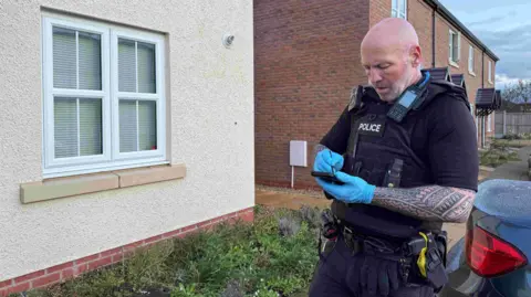 A police officer who has a shaved head and has tattoos stands in uniform marking details into his phone. He is wearing blue gloves and has several items of police equipment on his stab-proof vest. He is standing outside a suburban house. 