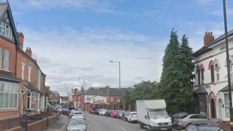 Google A road with terraced houses and parked cars on either side. The cars range in colour from white to red to black and there is also a white van in among them. The homes are made of brick with bay windows and small front gardens with brick walls at the front.