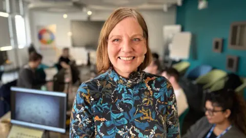 A woman wears a black top with a blue and orange leaf design. Behind her is a blue-walled room with many desks and screens, with six individuals occupying some of the desks. The wall behind her right shoulder is white with blind-covered windows. 