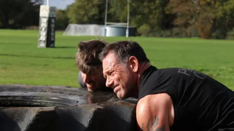 SSAFA/Sway PR Foxy wearing a black vest and training with a friend on a green field. They are both attempting to lift a huge tyre.