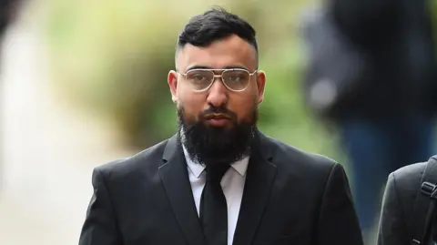 PA Media A young man wearing metal-framed glasses looks straight ahead and walks towards the camera wearing a black suit and tie and white shirt. 