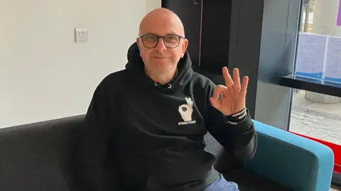 A man with a shaved head and black rimmed glasses sits on a small sofa. He is wearing a black hoodie with a logo showing a hand making the okay sign. He is making the same sign with his left hand.