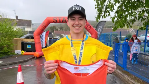 Chris Taylor smiles at the camera at the start line of the race. He is dressed in a yellow cheese costume and is wearing a black baseball cap. He is holding a red MND Association vest in both hands.