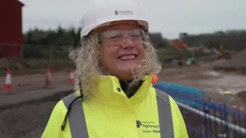 Lulu Bowerman smiling at the camera has blonde curly hair and is wearing a white hard hat and yellow hi-vis jacket.