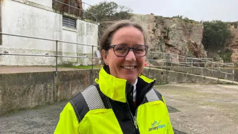 BBC Jeanette Sheldon wearing a hi vis jacket with the Jersey water logo on it. She has her hair tied up and is wearing glasses. Behind her is a white building and a cliff face. The sky is blue. 