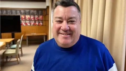 Kevin wearing a dark blue jumper in an office in the council building, smiling at the camera