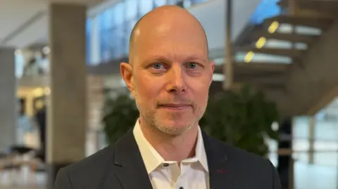 Marketing Director Ian Cook. He is standing in the large open reception area of the Cambridge University Press and Assessment building, wearing a suit.