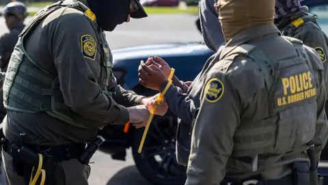 Two Border Patrol agents wearing green uniforms put a plastic tie around a man's hands. 