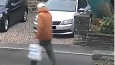 West Yorkshire Police CCTV image of a man in an orange jacket, carrying a plastic bag, walking along a residential road