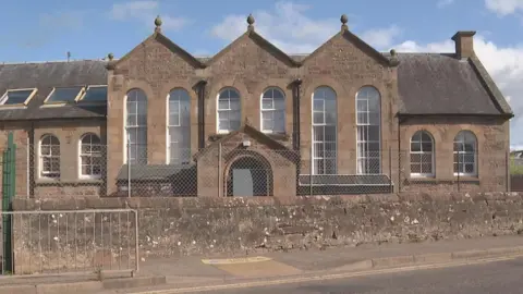 The school is from the Victoria era. It has long narrow windows, which are arch-shaped at the top. The school is behind a long stone wall.