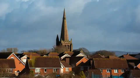 Millom Town Deal Board A church spire rises above several houses in Millom. Hills can be seen in the distance.

