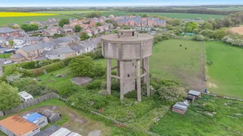 Savills An aerial photo of the water tower which is surrounded by fields and gardens and shows no road leading up to it. There are horses grazing in the field behind it on the right and a settlement of houses behind it on the left.
