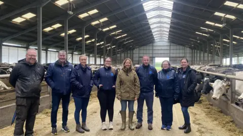 Marie Stephenson Marie Stephenson with a group of colleagues stood in a barn with cows in it.