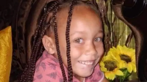 Joshlin Smith, wearing her hair in braids, smiles at the camera. Sunflowers can be seen behind her.
