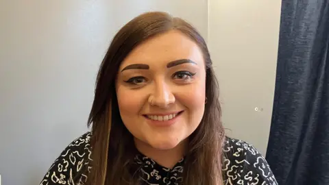 A brown haired woman in a patterned blouse sitting in a kitchen