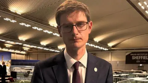 Simon Thake/BBC A man stands in a leisure centre where tables are in the background for the vote count. He has a navy suit, burgundy tie, brown short hair and black square glasses. He has a neutral expression but looks sad.