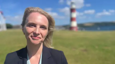 Charlotte Holloway wearing a mint top and navy blue blazer on Plymouth Hoe with Smeaton's Tower lighthouse in the background.