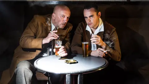 Eoin Carey/Citizens Theatre Two men, one bald, one with thick dark hair, sit side by side in 1950s costumes, drinking and smoking.