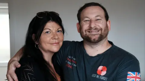 John Fairhall/BBC A head and shoulders image of a woman and a man, who has his arm around the woman. She is wearing a black top and has glasses on her head. She is smiling and looking into the camera. The man is wearing a T-shirt with a red poppy on it and the words Lest We Forget. He is smiling and looking into the camera.