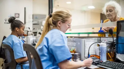 Stock photo shows two NHS receptionists sitting at a desk in front of computers behind a glass pane with a hospital visitor on the other side of the pane being attended to by one of the staff, in a hospital setting.