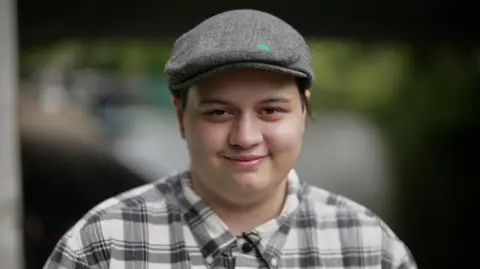 Ezra Quinton, wearing a grey flat cap with a green mark on it and a black and white checked shirt with a button down collar is looking straight into the camera and smiling, against a blurred greenish background.