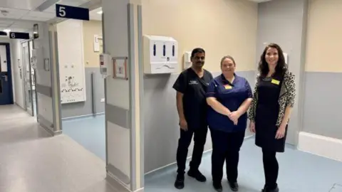 The Shrewsbury and Telford Hospital NHS Trust Three people stand in a hospital, in a large open cubicle. A female clinician stands in the middle, wearing trousers and a navy hospital shirt, a man on her left in black scrubs. A woman on the right with long brown hair wears a leopard-print cardigan.