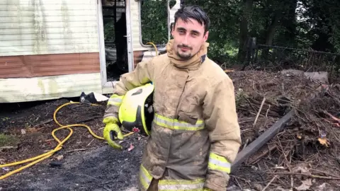 Oliver Dent Oliver Dent wearing a firefighter's uniform with a yellow helmet tucked under one arm and standing in front of a burnt caravan.