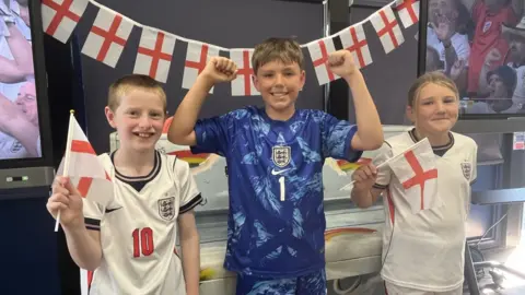 Three pupils stand smiling the the camera. They are wearing England football kits. 