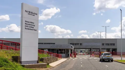 The entrance to Nissan's factory in Sunderland. A large sign stands outside the plant. A car drives towards the entrance of the long and low grey building. Railings and gates around the site are painted a vibrant red colour.