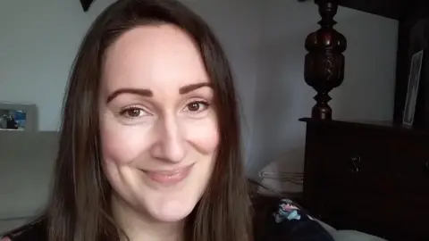 A woman with long dark hair takes a selfie at her home with a wooden chest and a beige sofa behind her.