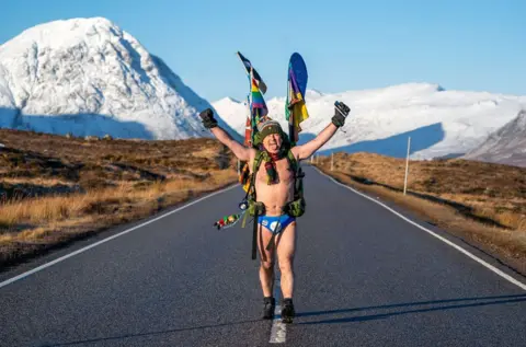 PA Media Mick Cullen, otherwise known as Speedo Mick, walks through Glencoe in sub-zero temperatures wearing only his swimming trunks. He is  wearing his blue Speedos, a woolly hat, scarf, backpack, gloves and nothing else, his face tilted upwards to the sky with his eyes closed and a look of relief, against a background of snowy mountains in Glencoe, Scotland, in January 2023
