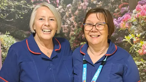 Two women are stood in front of a wall with images of flowers on it. The woman on the left has grey hair, the woman on the right has brown hair and is wearing glasses. Both are smiling and wearing navy blue nurses gowns.