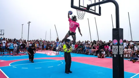 Sam Neter A person in a pink shirt jumping over a female police officer and dunking a basketball on a blue and pink court.