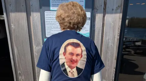 Sally Cooke A woman is standing with her back to the camera. She has a blue T-shirt on with a picture of her late husband.