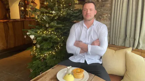 Olivia Richwald/BBC Tommy Banks, pictured in front of a Christmas tree with a plate of pie and mash in front of him. Mr Banks is wearing a white shirt and dark trousers and is sitting with his arms crossed.