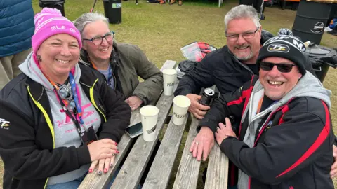 BBC Two couples in big coats and hat sit around a table and smile.