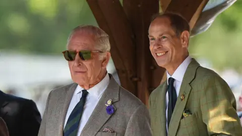 PA King Charles wearing a tweed blazer, a green and navy striped tie and brown Serengeti sunglasses. He is with a smiling Prince Edward who is wearing a green blazer.