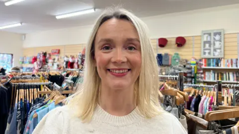 Jennifer Rowan smiles at the camera as she stands in Dove House shop. She has shoulder-length blonde hair and is wearing a cream crew kneck jumper.