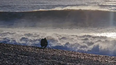 Sarah Giles A person crouching next to a wave on a pebble beach. The wave is bigger than the person.