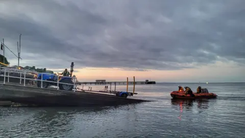 RNLI Penarth Lifeboat Station An orange RNLI lifeboat heading out to sea from a concrete slipway. Three RNLI members can be seen on the boat with helmets and life vests. A man sits on a blue tractor halfway up a ramp leading into the sea, he looks back at the lifeboat. The sun is setting in the background behind grey clouds, and Penarth Pier can be seen in the background. 