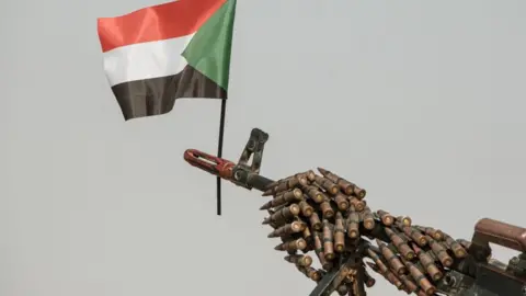 A Sudanese flag placed above the muzzle of a machine gun covered with an ammunition belt.