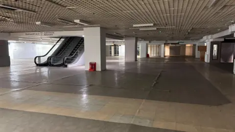 Jodie Halford/BBC A former shop floor has large, empty floorspace with an escalator on one side and columns supporting the ceiling above. The floor is concrete.
