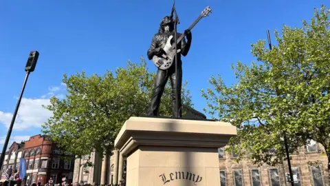 The cast bronze statue showing a man with a guitar in front of a microphone. It stands on a stone plinth with Lemmy written on it. There are buildings and trees surrounding it.