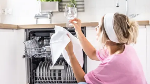Getty Images A young woman at home inspects a glass that has been washed in the dishwasher to a shine