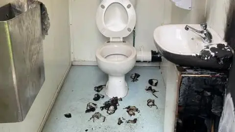 The single toilet cubicle has been vandalised. The sink on the right is blackened and torched from fire damage. There are burn marks on the blue floor and the white wall above the toilet is blackened also. There is a metal bin on the wall to the left.
